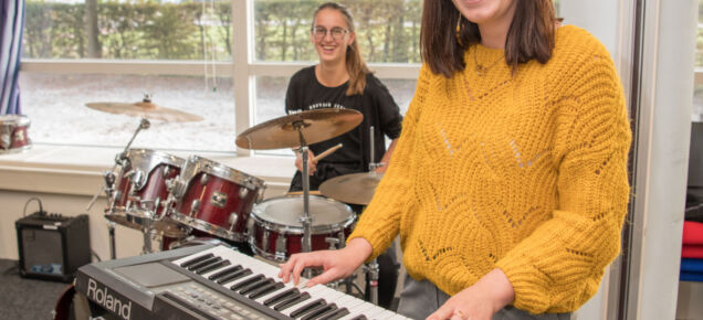 Een student die speelt op een keyboard en een drummende student op de achtergrond