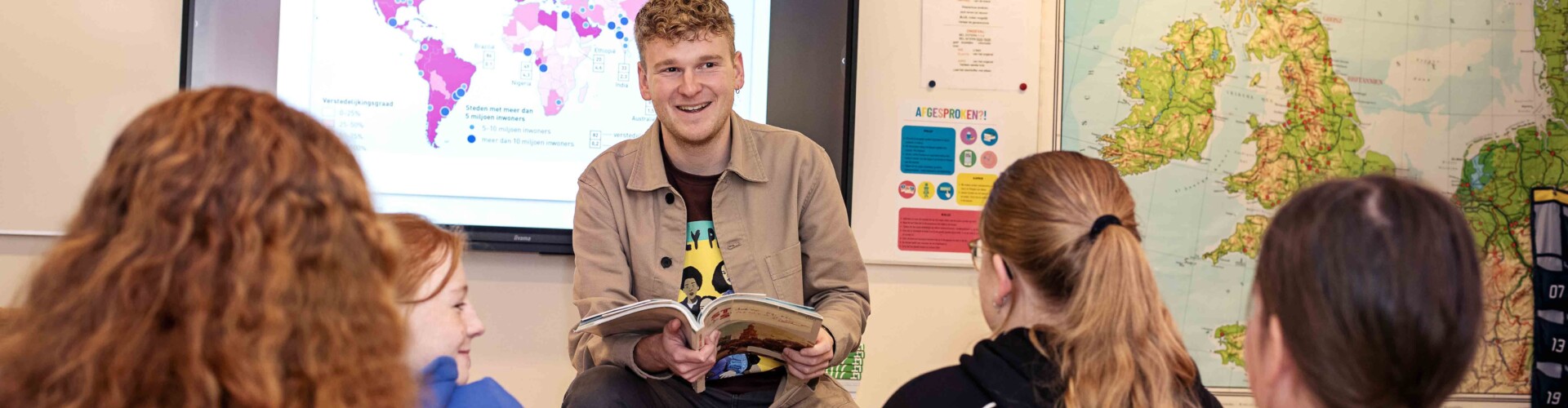 Een jonge docent die enthousiast lesgeeft aan een groep studenten in een klaslokaal. Op de achtergrond zijn een wereldkaart en een projectie van een wereldkaart met data zichtbaar.