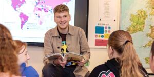 Een jonge docent die enthousiast lesgeeft aan een groep studenten in een klaslokaal. Op de achtergrond zijn een wereldkaart en een projectie van een wereldkaart met data zichtbaar.