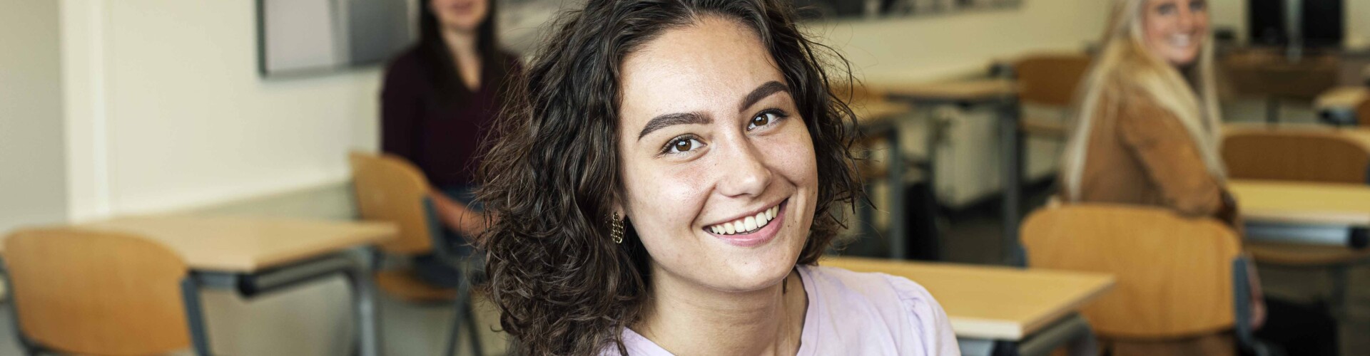 Pedagogiek student poseert in een klaslokaal met twee andere studenten op de achtergrond