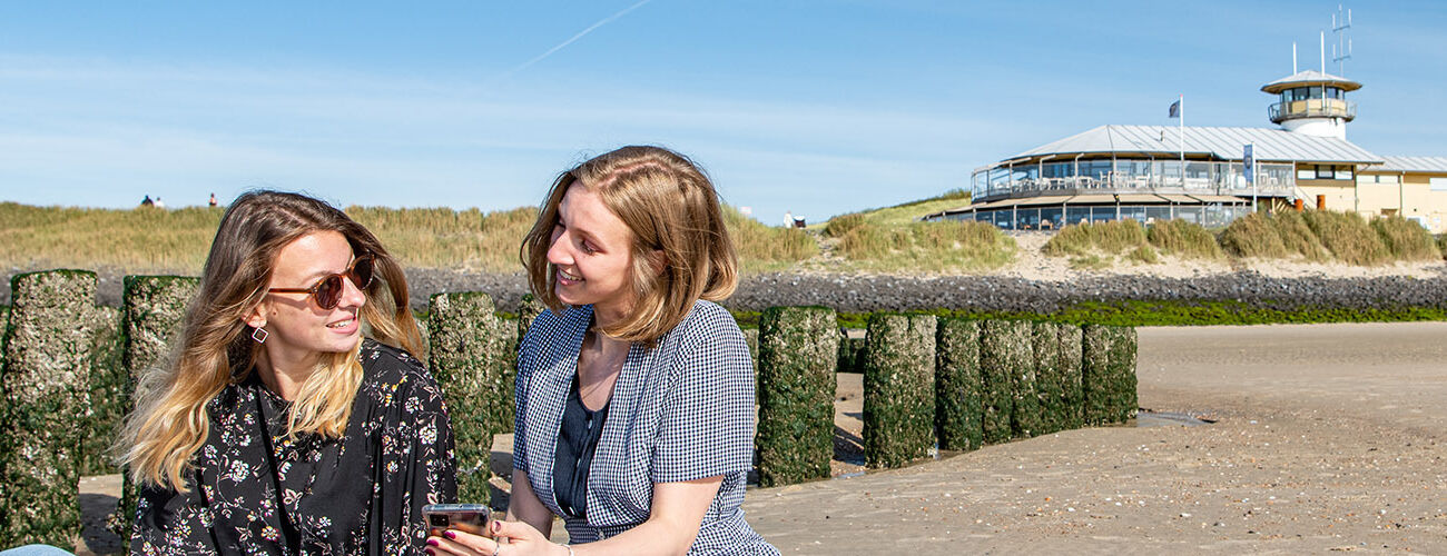 Two female international students are sitting at a typical 'Zeeuws' beach, located around Vlissingen, Zeeland, The Netherlands. One student shows the other student something on her phone and the smile at each other.