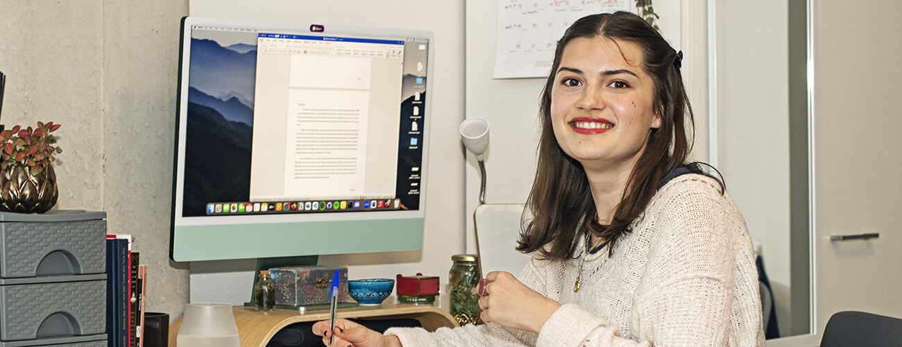 A young international student living alone while studying abroad in the Netherlands, is sitting at her desk in her room behind the computer working on a paper en looking in the camera.