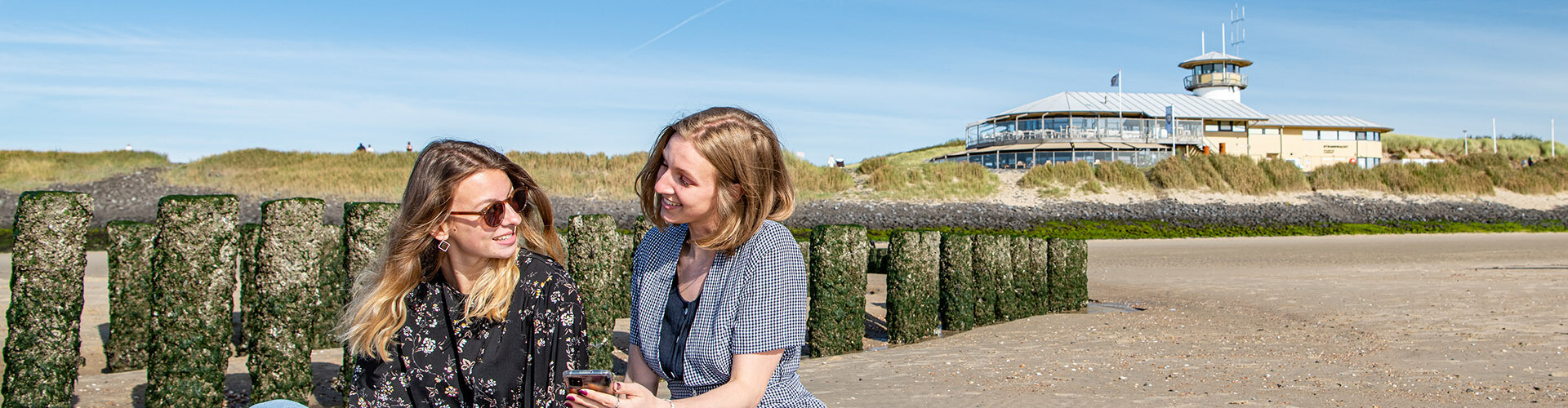 Two female international students are sitting at a typical 'Zeeuws' beach, located around Vlissingen, Zeeland, The Netherlands. One student shows the other student something on her phone and the smile at each other.