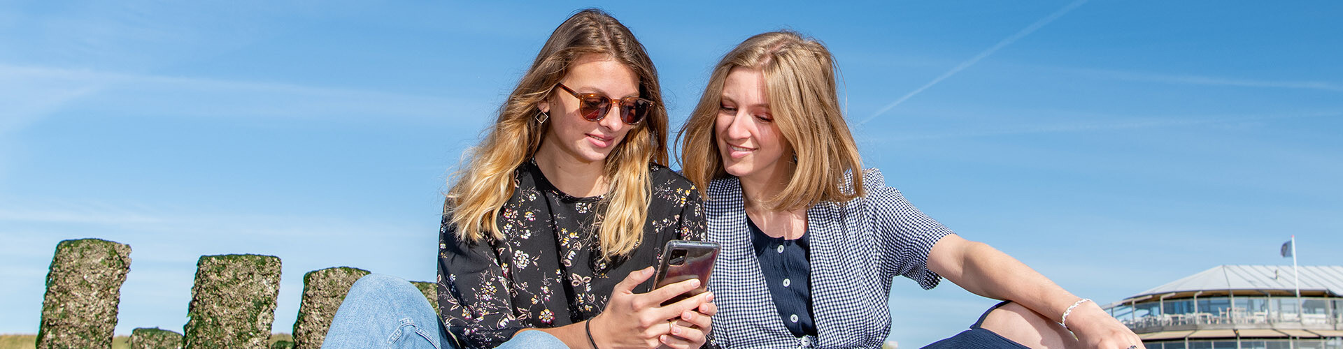 Two female international students sitting on the beach in Zeeland, the Netherlands, looking at student jobs on their phone.