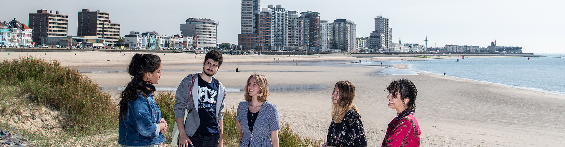 A group of international students, who study at HZ University of Applied Sciences, laughing an talking in the dunes of the beach at Vlissingen, Zeeland, The Netherlands, with the sky line of sea and the City of Vlissingen in the background.