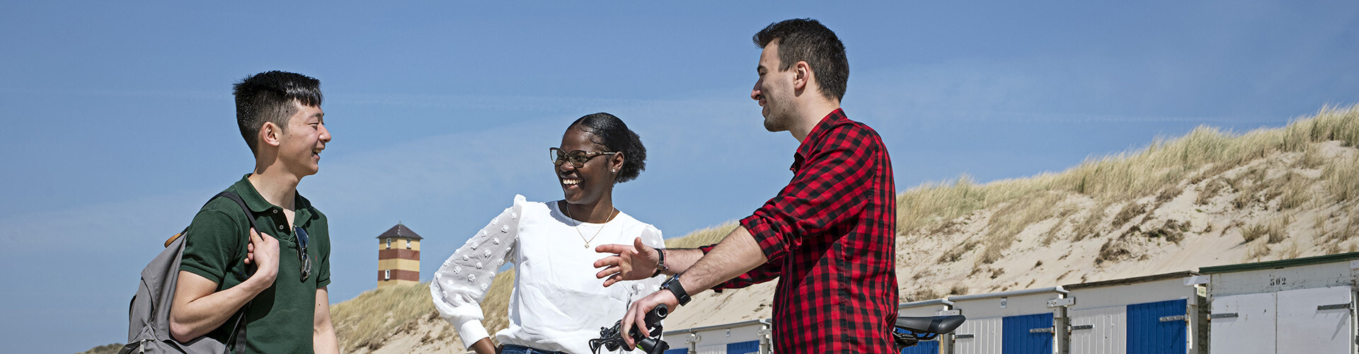 Three international students (one girl and two boys) are talking and enjoying themselves at the beach in Vlissingen, Zeeland, the Netherlands with the dunes and lighthouse in the background.