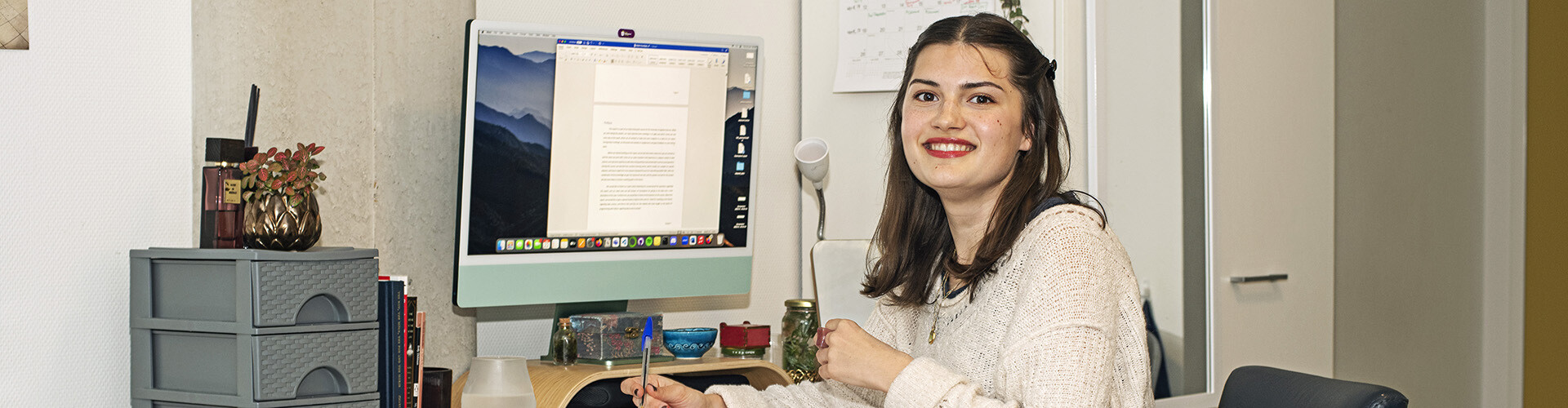 A young international student living alone while studying abroad in the Netherlands, is sitting at her desk in her room behind the computer working on a paper en looking in the camera.