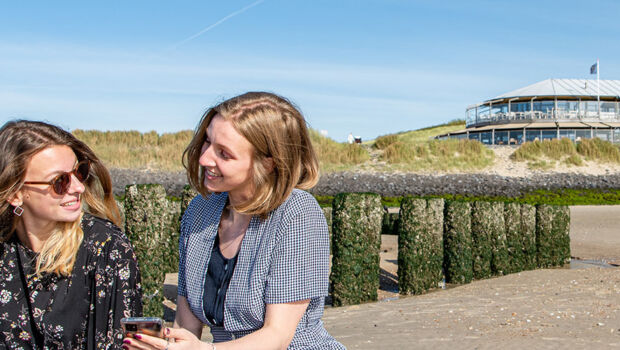 Two female international students are sitting at a typical 'Zeeuws' beach, located around Vlissingen, Zeeland, The Netherlands. One student shows the other student something on her phone and the smile at each other.