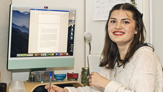 A young international student living alone while studying abroad in the Netherlands, is sitting at her desk in her room behind the computer working on a paper en looking in the camera.